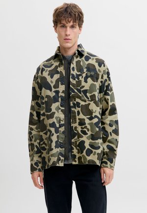 Joven con una camisa de camuflaje verde y marrón abotonada sobre una chaqueta negra con cremallera, de pie frente a un fondo liso y claro.