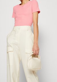 Top corto a maniche corte di colore rosa a coste abbinato a pantaloni cargo a gamba larga color crema. Una mano tiene una piccola borsa a mano di forma rotonda di colore crema.