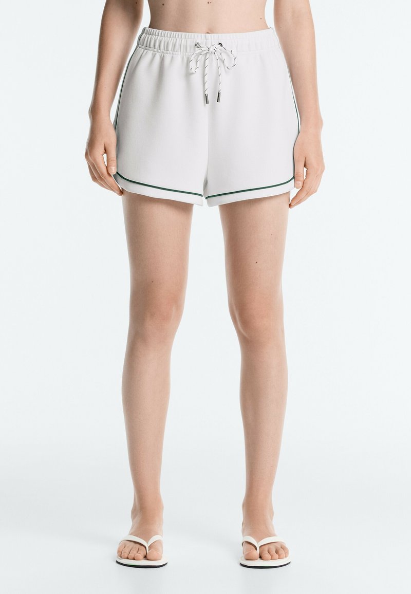 Personne portant un short blanc à taille élastique avec une bordure foncée et des tongs blanches, debout devant un fond clair uni.