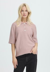 Różowy sweter z dzianiny z kołnierzykiem, krótkimi rękawami i trzema guzikami. Teksturowana tkanina z ribbowanymi brzegami, w parze z czarnymi spodniami.