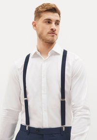 Jūrliniai petnešos pritvirtintos prie baltos marškinėlių, turinčių vertikalią gydymo tekstūrą ir sidabrinę aparatūrą, dėvimos ant mėlynų kelnių.