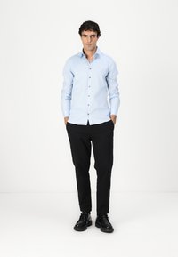 Lichtblauwe overhemdd met lange mouwen, bruine knopen, zwarte broek en zwarte schoenen. Eenvoudig ontwerp, slim fit, gladde stof.