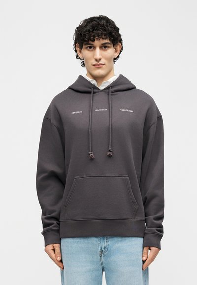 Jaunas žmogus su garbanotais plaukais, dėvintis tamsiai pilką gobtuvą su mažomis baltomis raidėmis ir šviesiai mėlynas džinsus, stovintis prieš šviesią foną.