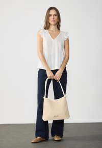 Biała, teksturowana bluzka z falbanistymi rękawami, w połączeniu z ciemnoniebieskimi dzwonami. Modelka trzyma beżową, plecioną torebkę z białymi ramiączkami.