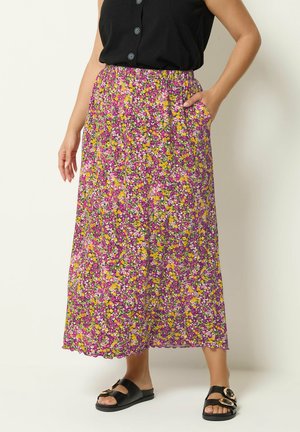 Femme portant une jupe midi fleurie rose, jaune et blanche, un top noir sans manches et des sandales noires à boucle, debout avec une main dans la poche.