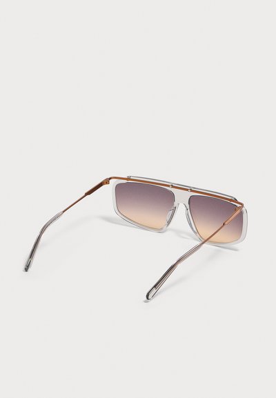 Lunettes de soleil rectangulaires avec verres dégradés du gris à l'ambre, fine monture métallique brune et branches en plastique transparent sur fond clair.