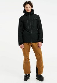 Schwarze wasserdichte Jacke mit zwei Brusttaschen, kombiniert mit braunen Skihosen und schwarzen Skischuhen. Glatte Textur, verstellbare Kapuze und Manschetten.