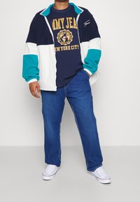 Niebiesko-biała kurtka na zamek z turkusowymi akcentami, nałożona na granatowy t-shirt z żółtym nadrukiem. Noszona z niebieskimi dżinsami i białymi trampkami.