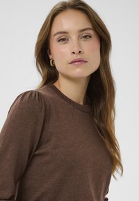 Rjava pletena majica z okroglim izrezom in napihnjenimi rokavi. Ima mehak teksturo in preprost dizajn, nosi pa jo z zlatimi obročastimi uhani.