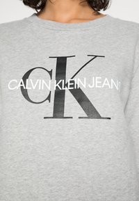 Šedá mikina s velkým logem. Černý text obsahuje "CK" a "CALVIN KLEIN JEANS" v kontrastní bílé, s hladkou strukturou.
