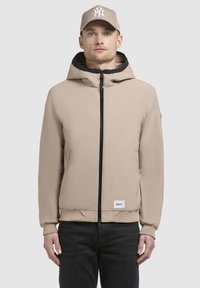 Giacca beige con zip, cappuccio bordato in nero, polsini a coste e un patch con logo sull'orlo, abbinata a jeans scuri e un berretto beige.