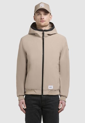 Beige zip-up jack met een zwart afgewerkte capuchon, geribde manchetten en een logo patch op de zoom, gecombineerd met donkere jeans en een beige pet.