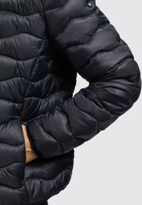 Osoba oblečená v čiernej prešívanej puffer bunde s rukou zasunutou do vrecka na jednoduchom svetlom pozadí.