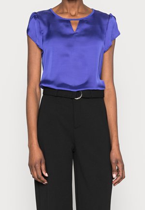 Žena nosí královsky modrou saténovou blůzu s krátkými rukávy a malým výstřihem vpředu, zastrčenou do vysokým pasem černých kalhot s páskem a stříbrnou přezkou.