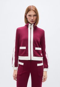 Μπορντό ζακέτα με φερμουάρ και λευκές λεπτομέρειες, με ψηλό γιακά, δύο μπροστινές τσέπες, ριμπ μανσέτες και ελαφρώς εφαρμοστή γραμμή.