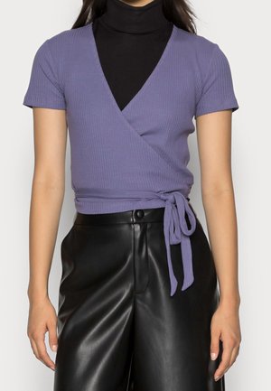 Fioletowa bluzka z prążkowanej tkaniny z krótkim rękawem i przewiązanym w pasie, nałożona na czarny golf, w zestawie z czarnymi skórzanymi spodniami.