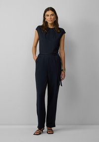 Salopette marine avec des manches courtes, taille ceinturée, coupe détendue et poches latérales. Associée à des talons noirs à brides et un bracelet métallique.