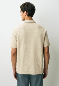 Chemise beige à manches courtes en tissu texturé avec broderie florale au dos. Design à col, ourlet droit.