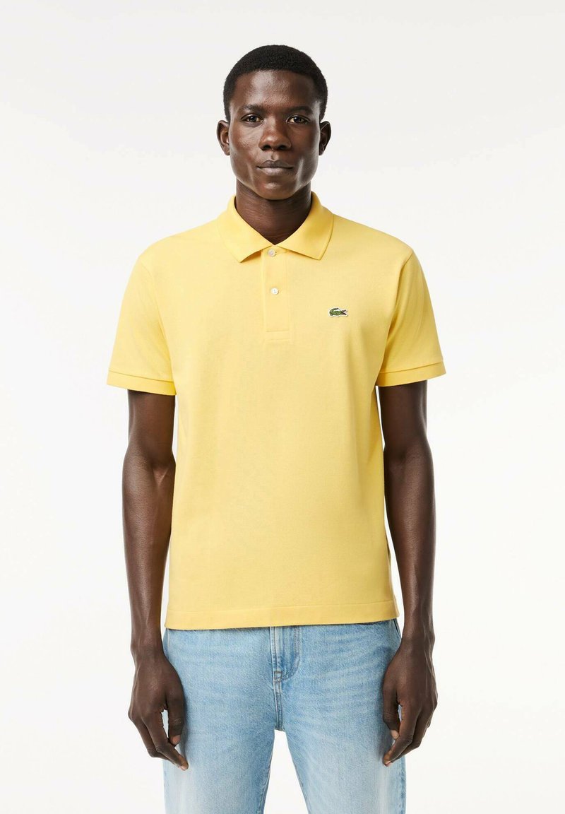 Camisa polo amarilla de algodón con un color clásico, dos botones y un logo de cocodrilo verde. Llevada con unos vaqueros de color azul claro.