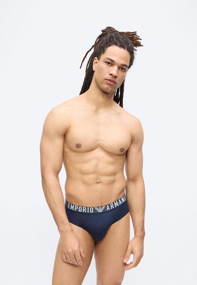 Άνδρας χωρίς πουκάμισο με ντρεντλοκς που φοράει σλιπ Emporio Armani σε ναυτικό μπλε χρώμα, πόζαρε σε απλό φωτεινό φόντο.