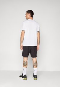 Witte T-shirt met korte mouwen gecombineerd met zwarte shorts. Toont tatoeages op de benen, draagt witte sokken en zwarte sportschoenen met groene accenten.
