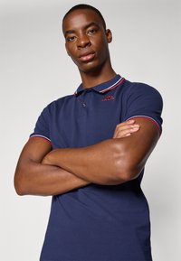 Mørkeblå polo skjorte med hvit og rød ribbet krage, med en liten brodert logo på brystet og kort erm. Glatt tekstur.