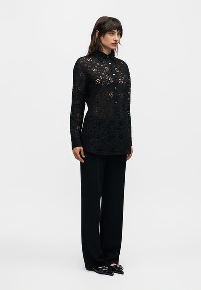 Γυναίκα που φοράει μαύρο μακρυμάνικο πουκάμισο με φλοράλ δαντέλα και κουμπιά, μαύρο παντελόνι με φαρδιά πόδια και μαύρα μυτερά παπούτσια με ασημένιες αγκράφες.