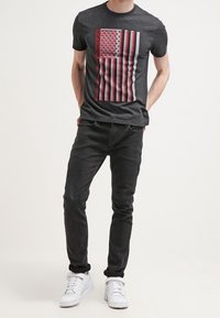Camiseta negra con un gráfico de un diseño de bandera en rojo, blanco y azul, complementada con jeans negros y zapatillas blancas.