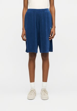 Person iført marineblå terry fabric shorts til knæet, hvide sokker, beige sneakers og en hvid ærmeløs top, stående på en ensfarvet baggrund.