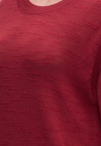 Gros plan d'un pull en tricot rouge texturé montrant son motif de tissu et son encolure arrondie sur une personne.