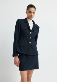 Blazer de raya diplomática en azul marino con botones dorados, dos bolsillos frontales, un dobladillo trasero alargado y una falda lápiz a juego. Acabado en tejido texturizado.