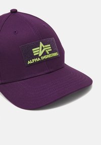 Textilből készült lila sapka, melyen egy texturált fekete folt található, zöld "ALPHA INDUSTRIES" logóval és egy háromszögletű emblémával.