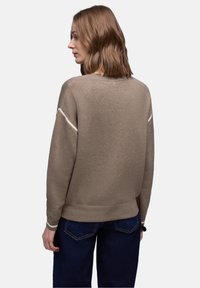 Femme aux cheveux mi-longs portant un pull beige avec des rayures blanches sur les manches et un jean bleu foncé, vue de dos sur un fond uni.
