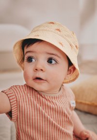 Chapeau bob jaune avec imprimé soleil, associé à une chemise à manches courtes rayée aux tons chauds. Texture douce, forme ronde, conçu pour le confort.