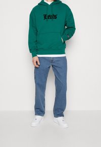 Grüner Baumwoll-Hoodie mit einer Kängurutasche und einem verstellbaren Kapuzenzug, versehen mit einem schwarzen Logo. Kombiniert mit blauen Jeans und weißen Sneakers.
