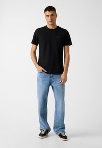 Schwarzes T-Shirt mit kurzen Ärmeln und Rundhalsausschnitt, kombiniert mit hellblauen, gerade geschnittenen Jeans und schwarzen Turnschuhen. Einfaches und legeres Design.