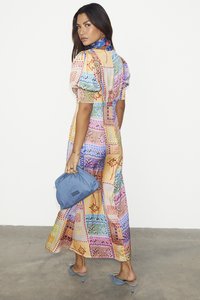 Robe maxi en patchwork multicolore avec des manches bouffantes, associée à une pochette bleue et des talons assortis, sur un sol en béton texturé.