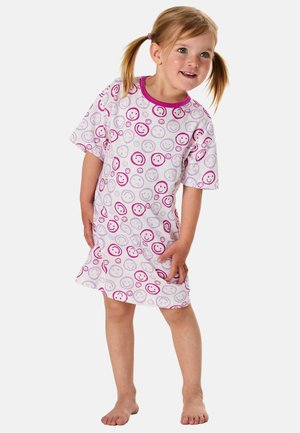 Junges Mädchen mit blonden Zöpfen, das ein weißes Kleid mit pinken und lila Mustern von lächelnden Gesichtern trägt und barfuß steht.