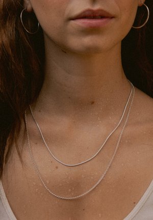Silberne Halsketten, übereinander getragen auf nackter Haut, mit glatten und strukturierten Gliedern, wobei eine Kette dicker zu sein scheint als die andere.
