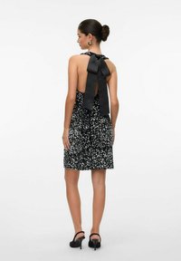 Juodų blizgučių suknelės galinis vaizdas su halter tipo kaklo ir dideliu juodu kaspinu. Audinys turi blizgantį tekstūrą ir prigludusią siluetą.