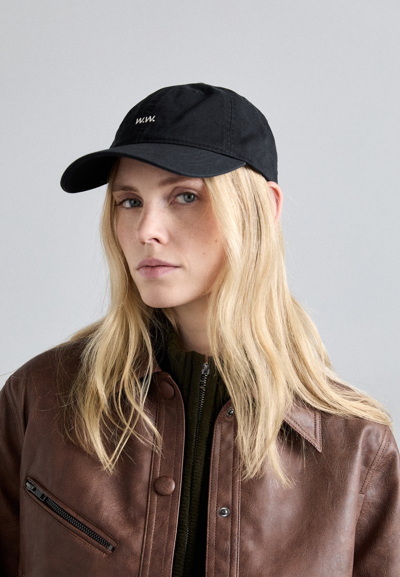 Svart baseballcap med brodert logo, med en buet skjerm. Brukt med en brun lærjakke og en grønn genser under.