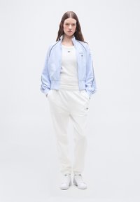 Γυναίκα όρθια με τα χέρια στις τσέπες, φοράει ανοιχτό μπλε ζακέτα με φερμουάρ, λευκή μπλούζα, λευκή φόρμα και λευκά αθλητικά παπούτσια σε απλό φόντο.