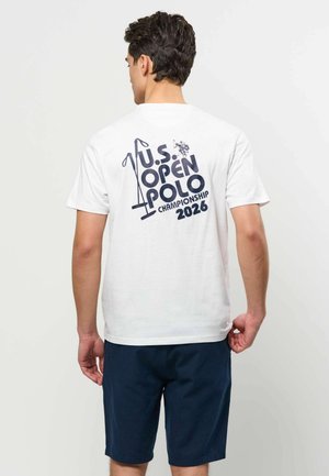 Mees, kes kannab valget T-särki tumelilla teksti ja graafikuga "U.S. Open Polo Championship 2026" seljal, tumelillad lühikesed püksid, seisab selgpoole vaatajale.