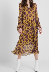 Dlouhé rukávy, žluté květinové šaty s fialovými vzory, výstřih do V, splývavá látka a volánkový lem. Nosené s bílými kotníkovými botami.