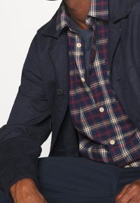 Giacca in cotone navy sopra una camicia a quadri rossa, blu e crema. Caratterizzata da chiusura con bottoni e una vestibilità rilassata con una finitura testurizzata.