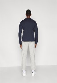 Άνδρας ντυμένος με μπλε μακρυμάνικο πουκάμισο, ανοιχτό γκρι παντελόνι φόρμας, λευκές κάλτσες και λευκά αθλητικά παπούτσια, στέκεται στραμμένος προς έναν λευκό τοίχο.