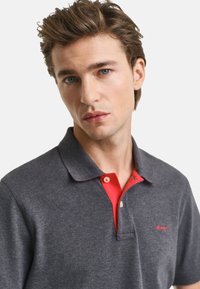 Charcoal-graues Polo-Shirt mit rotem Kragen. Verfügt über drei Knöpfe am Hals und ein kleines rotes Logo auf der linken Seite. Weiche Textur.
