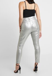 Pantalones ajustados metálicos plateados con cintura alta, que cuentan con cinco bolsillos y detalles de costura; combinados con sandalias negras de tacón alto.