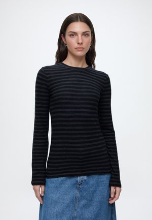Femme aux longs cheveux bruns portant un top à manches longues rayé noir et gris foncé et une jupe en jean bleue, debout devant un fond clair uni.