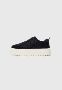 Adidași negri din suede cu cusături decorative, șireturi negre și talpă din cauciuc alb, cu un model ondulat, pe un fundal simplu deschis.